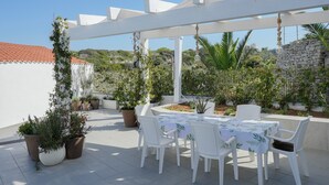 Un patio techado al aire libre con sillas y una mesa blancos, rodeado de plantas en macetas y una pérgola blanca.