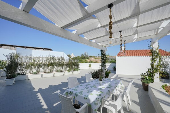 A rooftop terrace with a table and chairs, potted plants, and a clear view of the surrounding buildings.
