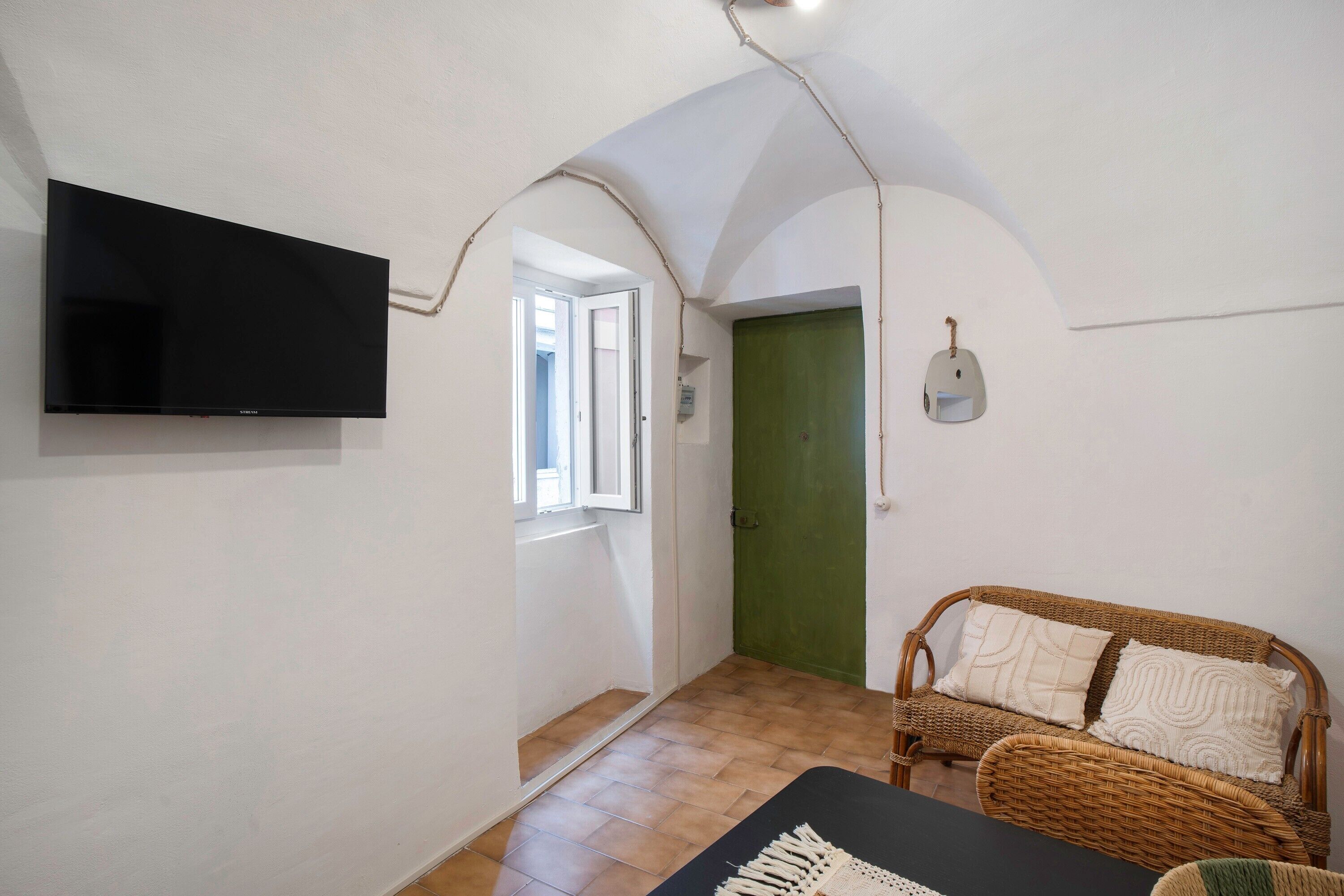 Una habitación con televisión de pantalla plana, un sofá de mimbre con cojines, una puerta verde y suelo de baldosas.