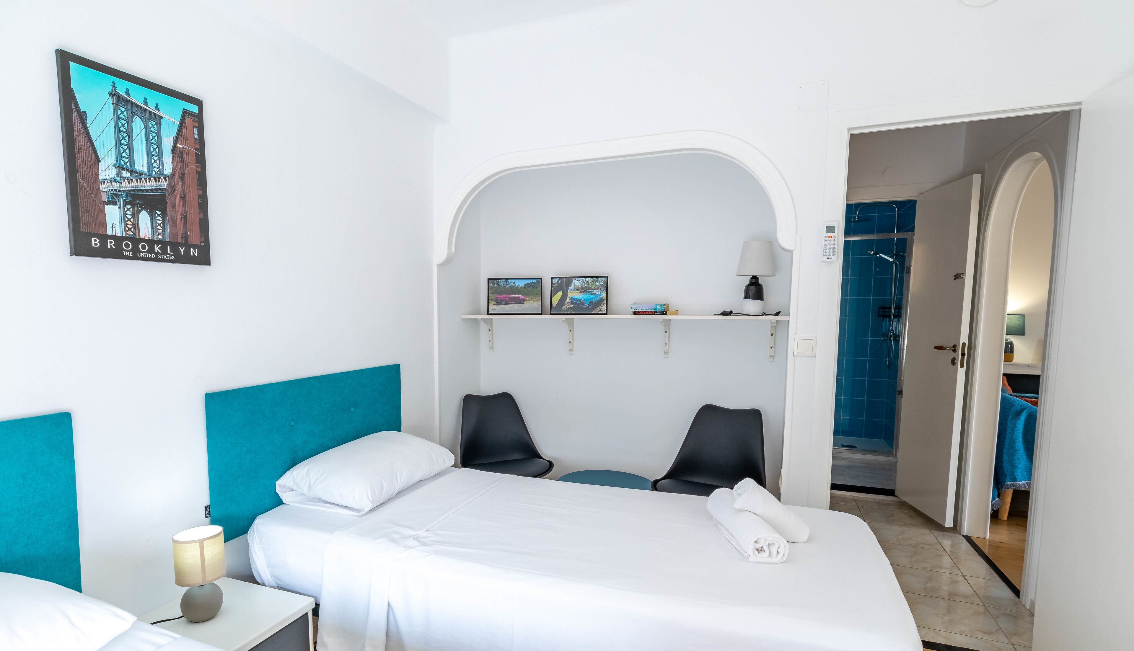 Een hotelkamer met een bed, twee stoelen, een nachtkastje, een lamp, een plank met foto’s en een deur naar een badkamer.
