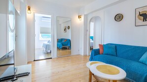 A modern living room with a blue sofa, a small white table, and a TV. There is a mirror reflecting the room and a door leading to another room. - Inftour Garvimar Apartamento (Calp)
