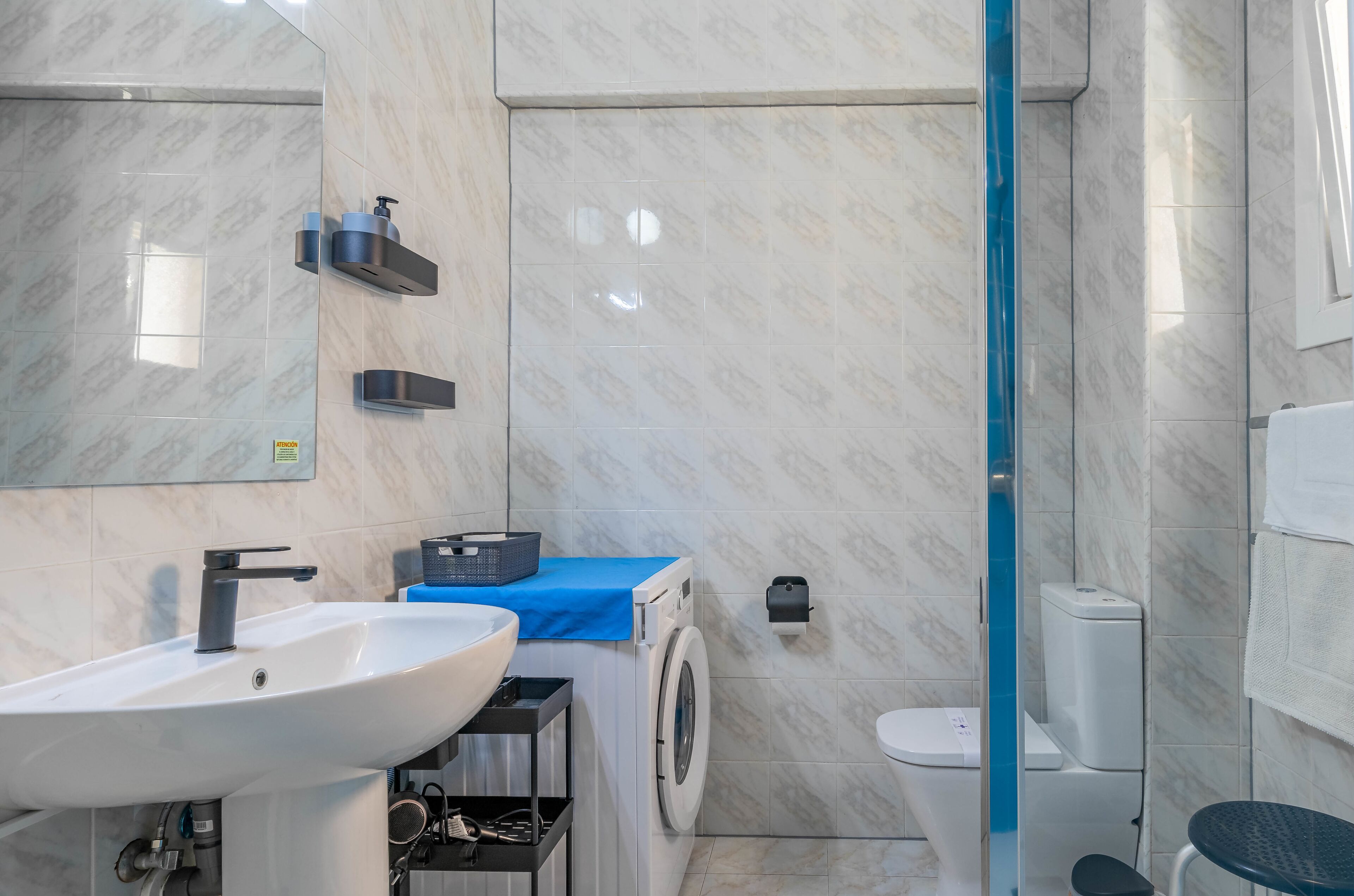 A modern bathroom with a sink, washing machine, toilet, and a glass shower enclosure.