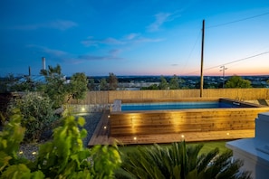 A swimming pool with wooden decking, surrounded by greenery and a stone wall.