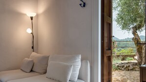 Un canapé avec des coussins, un luminaire fixé au mur et une vue sur un espace extérieur avec un arbre et une clôture.