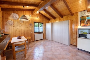 Interior de madera con una mesa de comedor, sillas, una lámpara colgante, una ventana y una zona de cocina con armarios y un microondas.