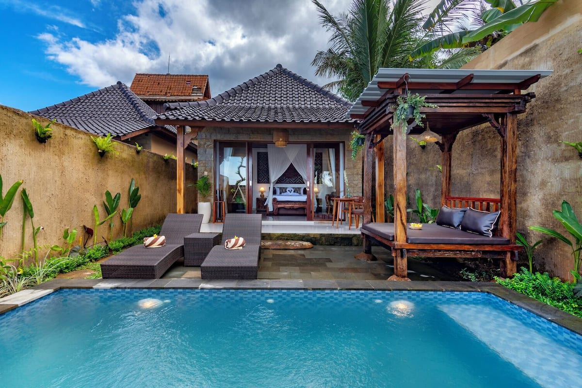 A poolside area with lounge chairs, a wooden pergola, and a view of a bedroom with a bed and curtains.