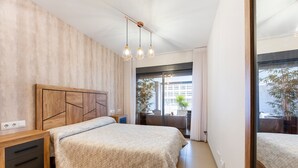 A bedroom with a wooden headboard, a bed with a textured blanket, a bedside table, a mirror, and a view of a balcony with plants.