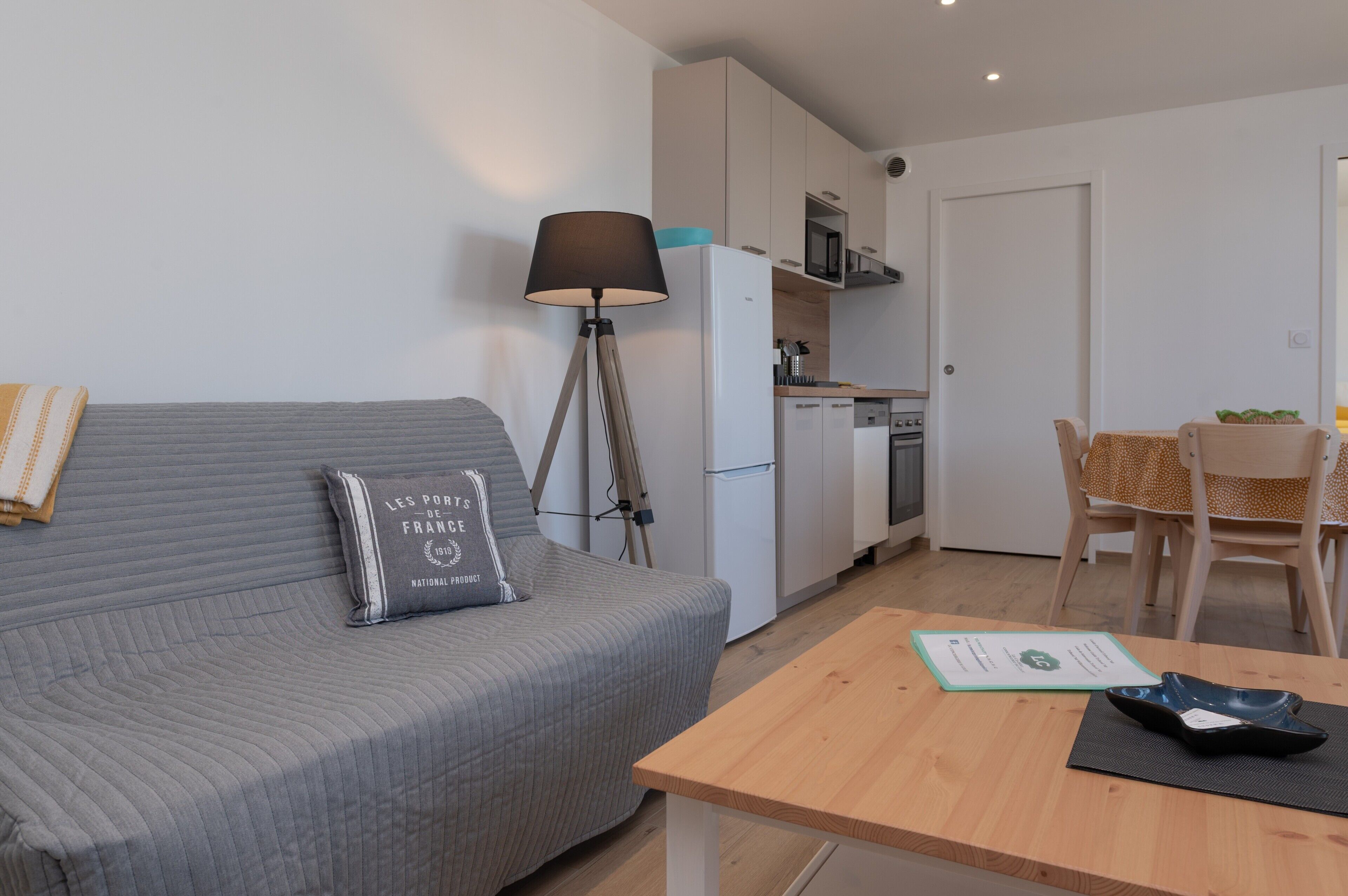 Ein modernes Wohnzimmer mit grauem Sofa, einer Lampe, einem Esstisch und einer Küchenzeile mit Kühlschrank und Mikrowelle.