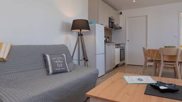 Ein modernes Wohnzimmer mit grauem Sofa, einer Lampe, einem Esstisch und einer Küchenzeile mit Kühlschrank und Mikrowelle.