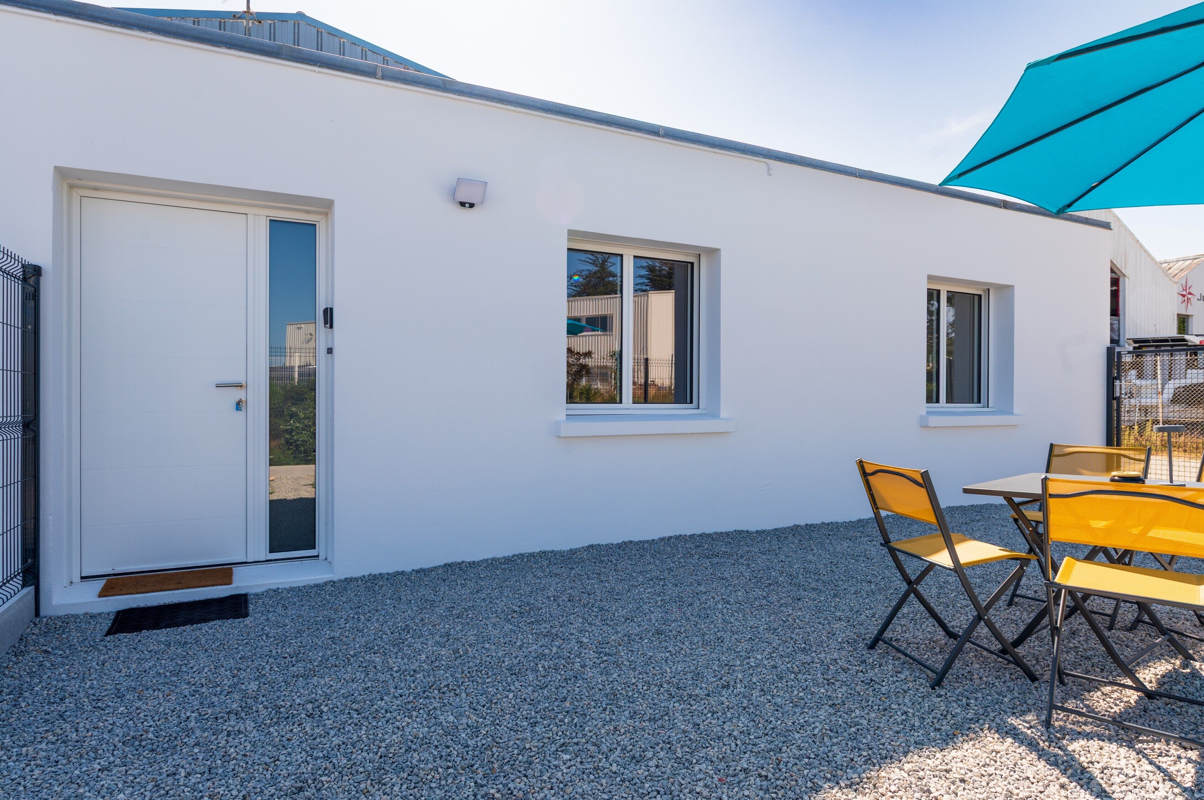 Un bâtiment blanc avec une porte, deux fenêtres et une terrasse en gravier agrémentée de chaises jaunes.