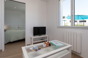 Una habitación de hotel moderna con una cama, un televisor sobre un soporte blanco, una mesa de vidrio con folletos y un ventanal con vistas al exterior.