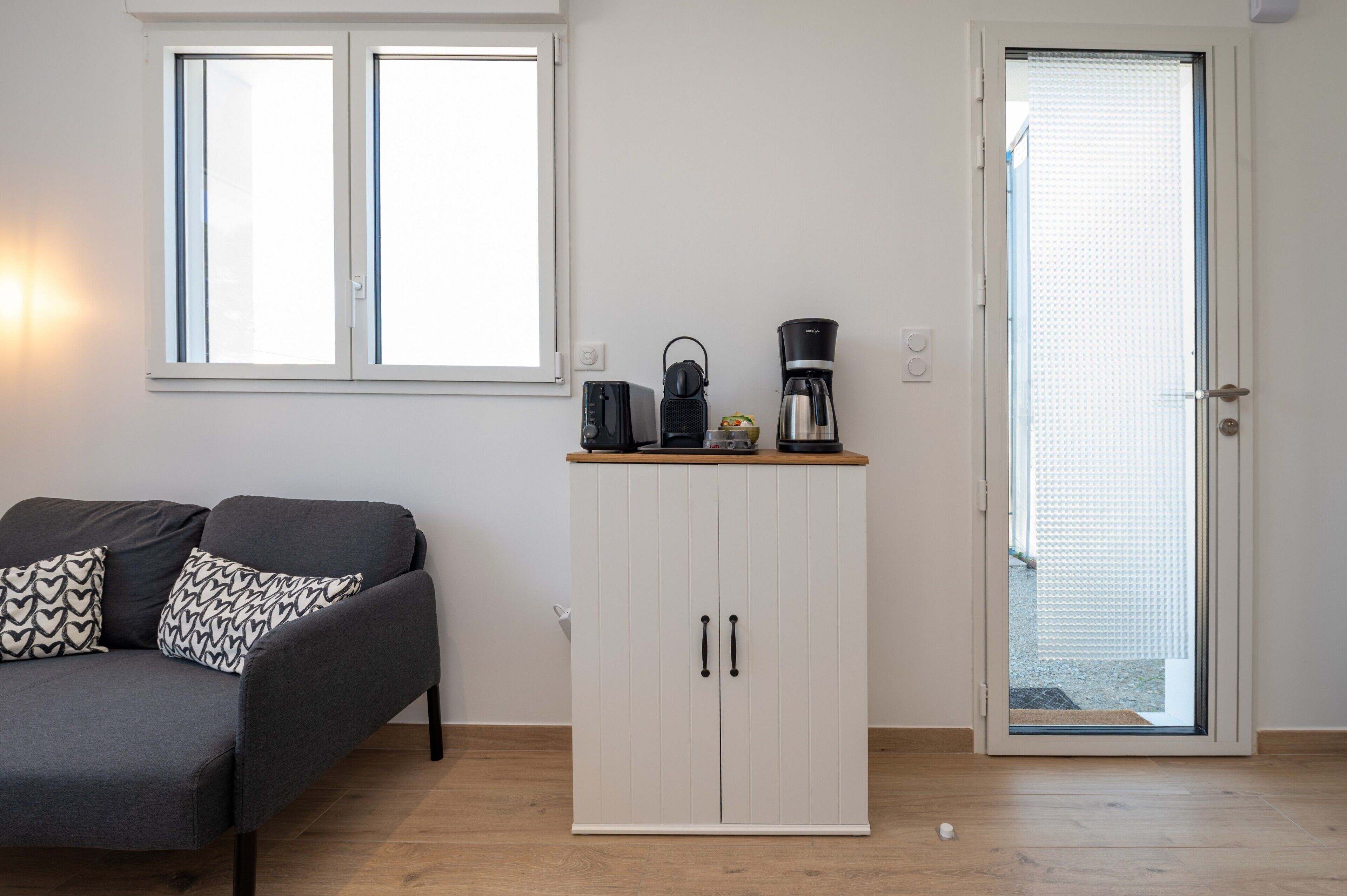 A room with a grey sofa, a white cabinet, a coffee maker, and a toaster on the counter.