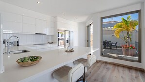 Une cuisine moderne avec des meubles blancs, un grand îlot central doté de tabourets de bar et une vue sur un patio agrémenté d’un palmier.