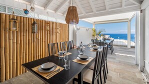 Un coin repas avec une table dressée pour quatre personnes, un mur en bambou, des lanternes suspendues et une vue sur l’océan.