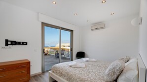 Une chambre à coucher comprenant un lit, une commode en bois, une porte-fenêtre coulissante donnant sur un balcon et un appareil de climatisation.
