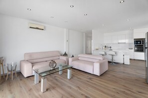 En moderne stue med et glasbord, en sofa og et køkkenområde med hvide skabe og hårde hvidevarer.