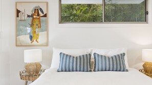 Un dormitorio con cama, dos almohadas, dos mesitas de noche, una lámpara, un cuadro y una ventana con vistas a zonas verdes.