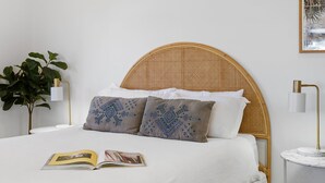 Ein ordentlich bezogenes Bett mit einem gewobenen Kopfteil, zwei Kissen mit blauen Mustern, einer weißen Decke und einem Buch darauf.