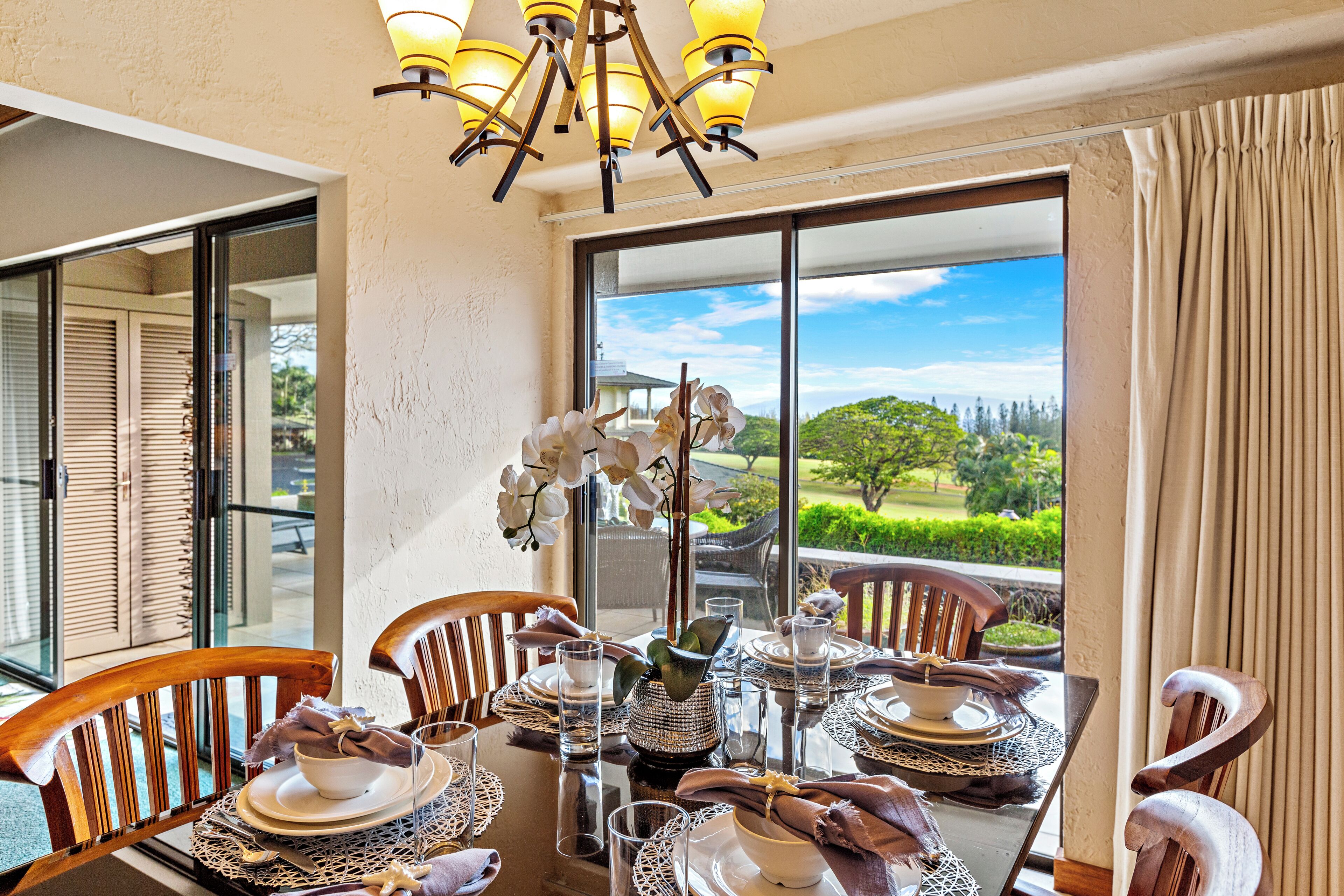 Comedor con una mesa dispuesta para cuatro, ventanas amplias con vista a zonas verdes y una araña.