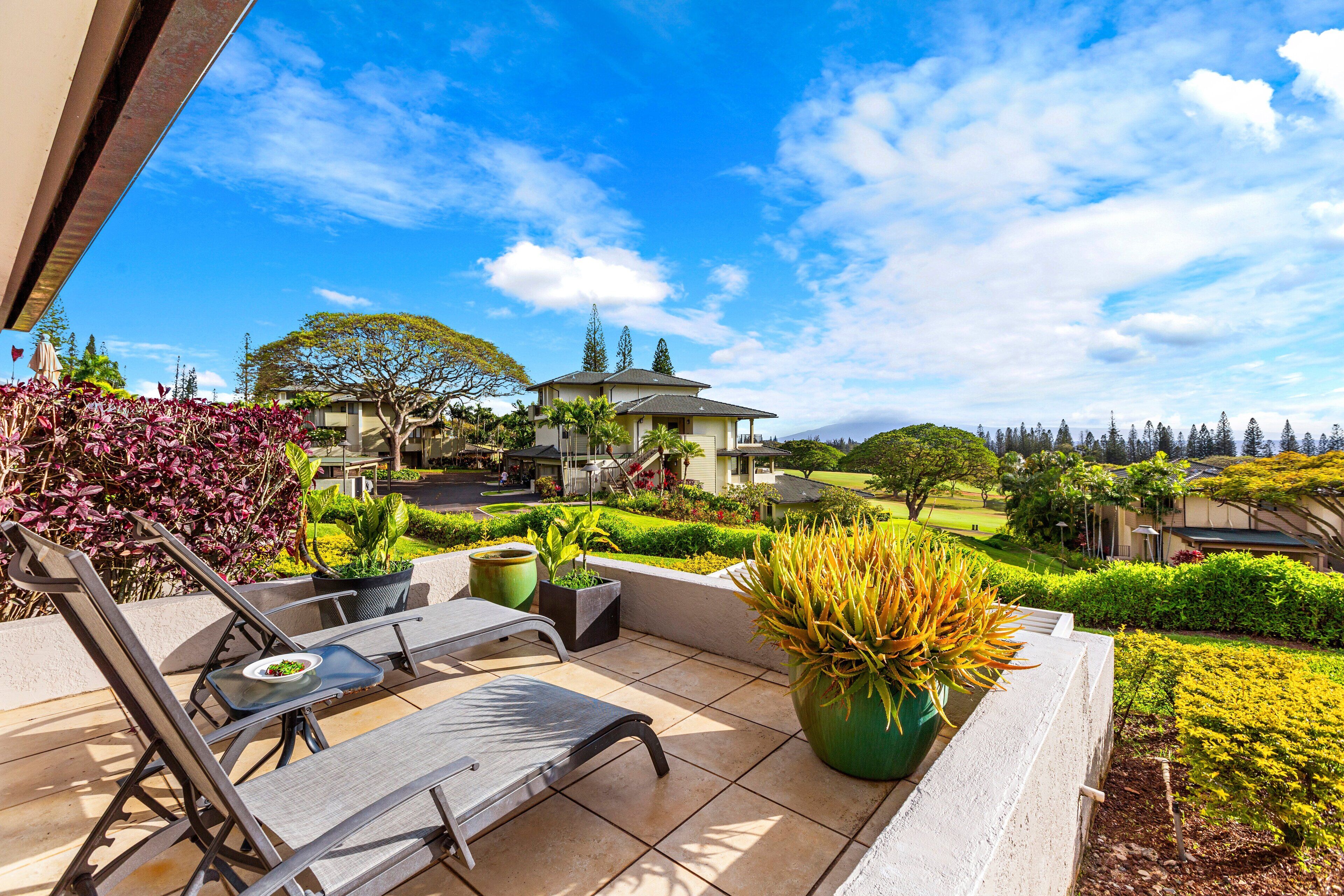 Une terrasse avec deux chaises longues et une table, entourée de plantes en pot, et offrant une vue sur un terrain de golf et des bâtiments au loin.
