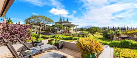 A patio with two lounge chairs and a table, surrounded by potted plants and a view of a golf course and buildings in the distance.