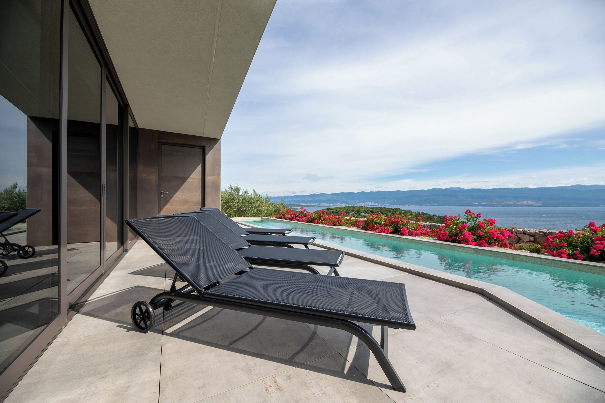 A modern outdoor pool area with lounge chairs, a clear view of the sea, and a stone wall with flowers.