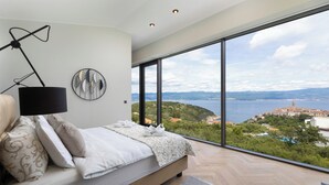 Een slaapkamer met een groot raam, een bed met wit beddengoed, een zwarte vloerlamp en een kunstwerk aan de muur.