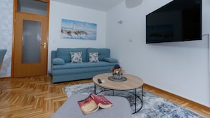 A modern living room with a blue sofa, a flat-screen TV, a wooden coffee table, and a painting of a lighthouse. - Trogir Stylish Apartment With Balcony and Sea View (Trogir)
