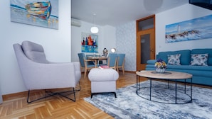 A modern living room with a grey armchair, a round coffee table, a blue sofa, and a dining area with wooden chairs and a table. - Trogir Stylish Apartment With Balcony and Sea View (Trogir)