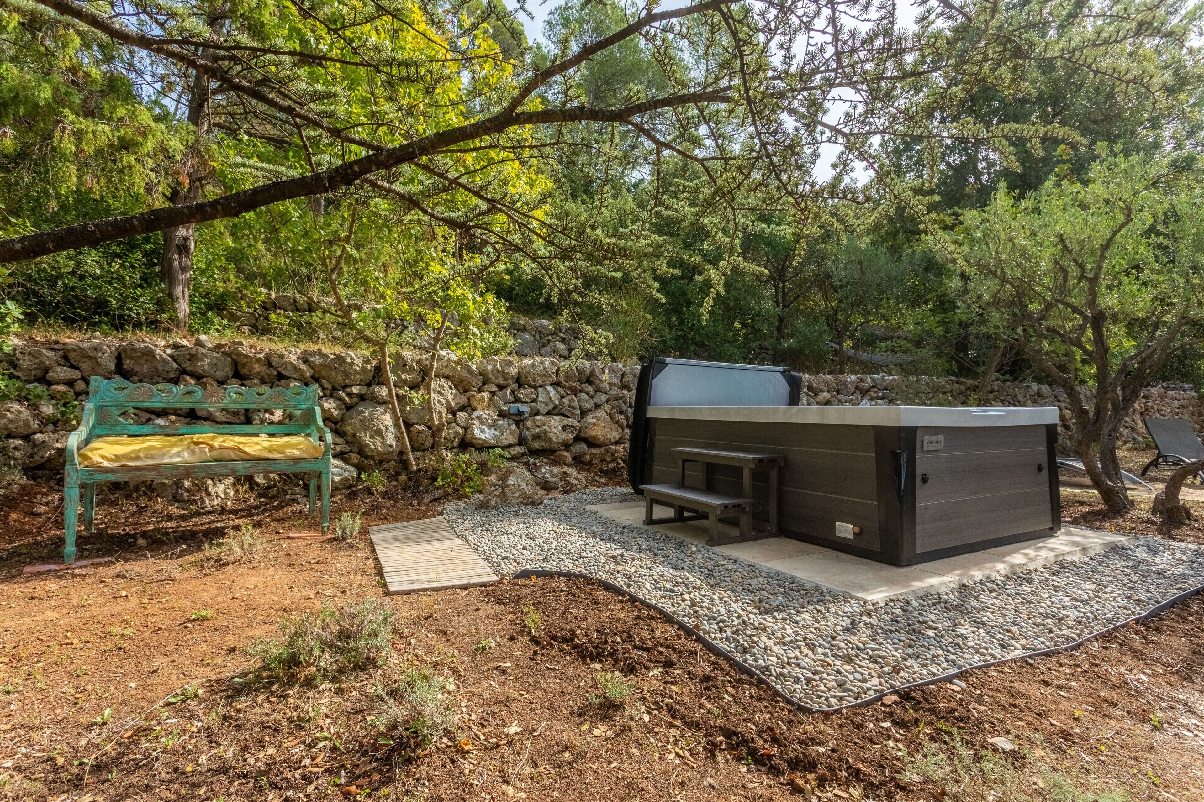 A stone wall, a bench, a hot tub, and a wooden deck in a forested area.