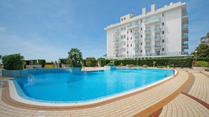 A swimming pool with a tiled deck, surrounded by greenery and a white building with balconies. - Oasis 38: Pool, Private Parking, Steps from the Beach (Lido di Jesolo)