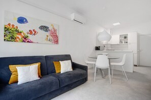 En moderne stue med en grå sofa, hvide og sennepsfarvede puder, et spiseområde med et hvidt bord og stole, samt et køkken med hvide skabe og hårde hvidevarer.