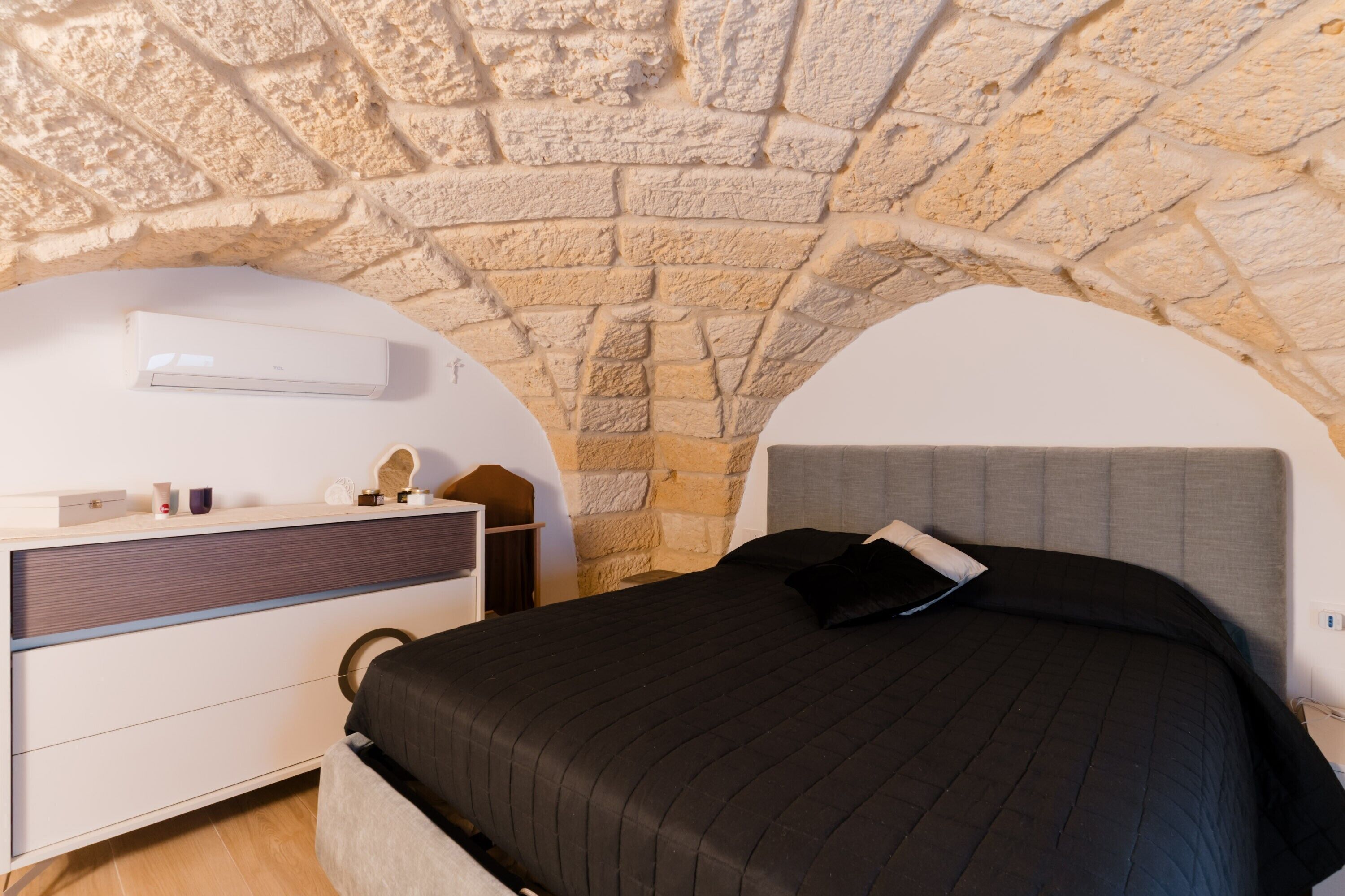 A bedroom with a stone arch ceiling, a bed with a dark headboard, a bedside table with a lamp, and a wall-mounted air conditioner.