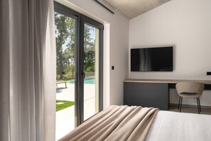 Quarto de hotel moderno com cama, escrivaninha, cadeira, TV e porta de vidro deslizante que dá para uma área de piscina.