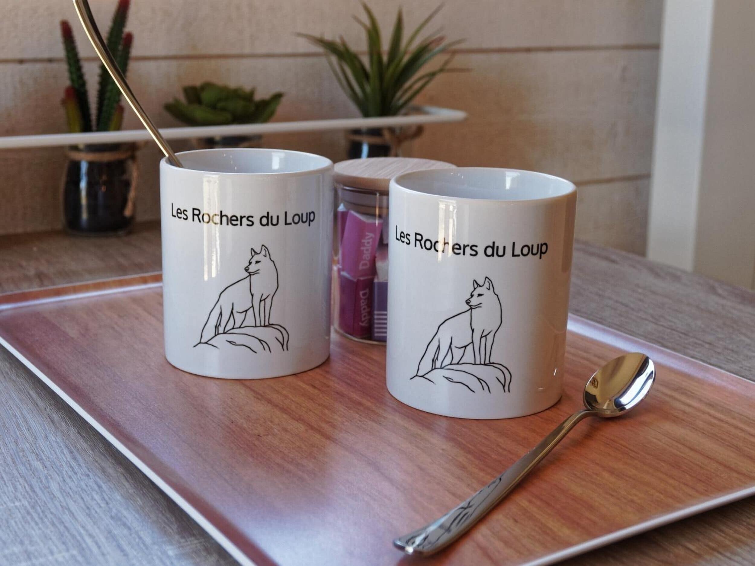 Two white mugs with a wolf illustration and text, placed on a tray with a spoon.