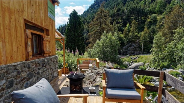 Eine Holzterrasse mit zwei Liegestühlen und einer Feuerstelle, umgeben von einer Steinmauer und einem Holzzaun, mit Blick auf ein bewaldetes Tal.