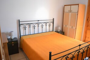 Une chambre à coucher avec un lit en métal, une couvre-lit orange, une table de chevet avec une lampe et une armoire.