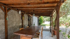 A wooden table with benches under a pergola, surrounded by stone walls and potted plants.