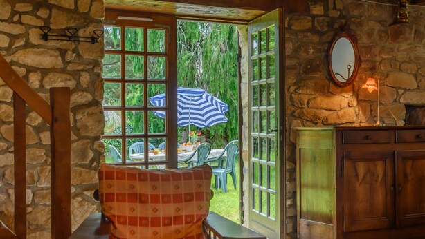 Une pièce rustique avec un mur en pierre, des poutres en bois, une table à manger avec des chaises et une vue sur la verdure à l’extérieur.