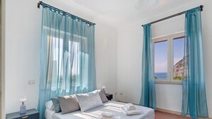 Quarto com cama, mesa de cabeceira, janela com cortinas e vista para o mar.