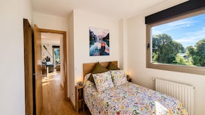 A bedroom with a bed, a window with a view, a painting on the wall, and a wooden door leading to another room.