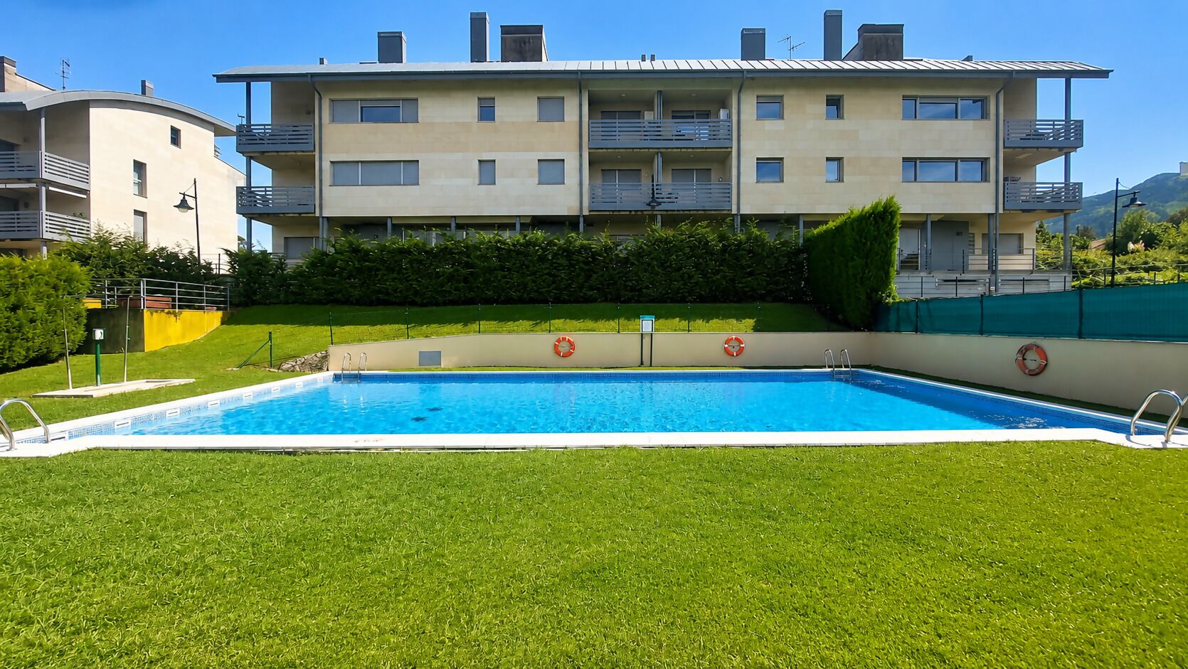 Una piscina rectangular con superficie de agua azul cristalina, rodeada de césped verde y bien cuidado. En el fondo, un edificio residencial de varios pisos con balcones y techo plano.
