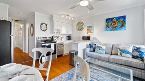 Une cuisine moderne avec des meubles blancs, un coin repas avec une table dressée pour deux personnes, et un salon avec un canapé et des coussins décoratifs.