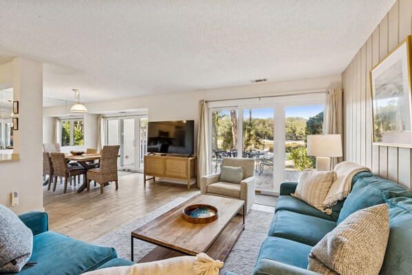 A modern living room with a sofa, coffee table, TV, and dining area with chairs and a table. - NEWLY LISTED! Spacious Beach Wood Villa Across from Beach Access! (Amelia Island)