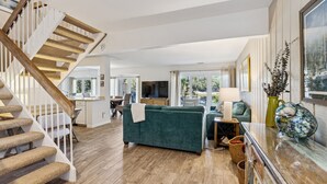 A spacious living area with a staircase, a green sofa, a dining table, and a kitchen in the background.