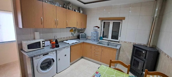 A compact kitchen with wooden cabinets, a washing machine, a microwave, a small refrigerator, and a window.
