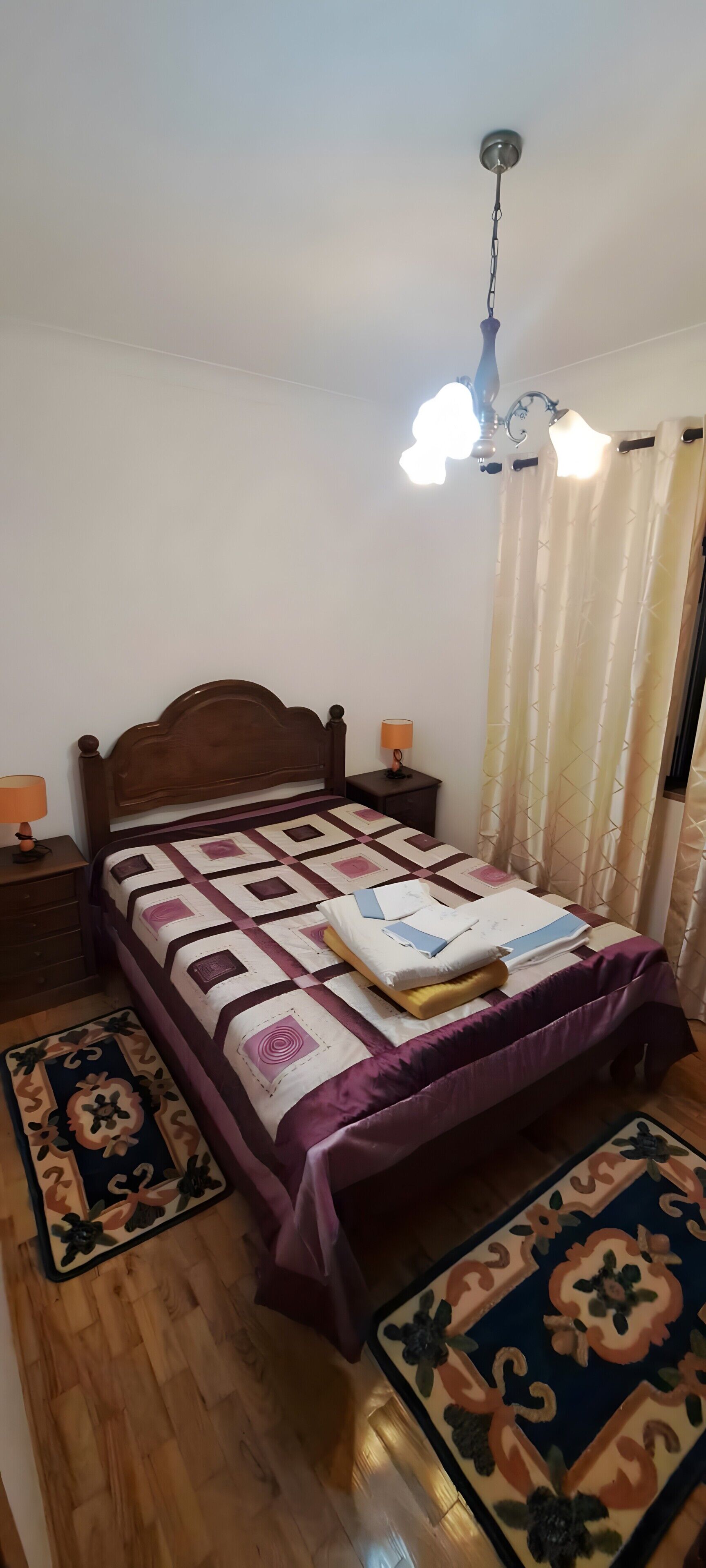 Een slaapkamer met een houten bed, een nachtkastje met een lamp, een plafondlamp en een kamerkleed met een patroon.