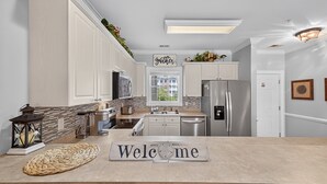A modern kitchen with white cabinetry, a stone backsplash, and a welcome sign.