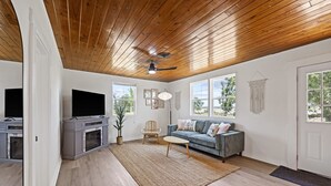 A spacious living room with a wooden ceiling, a gray entertainment unit, a blue sofa, and a large window.