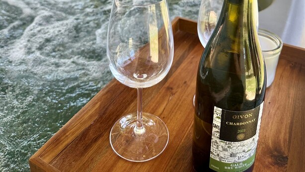 A hot tub with a wooden deck, a bottle of Givon Chardonnay 2023, and a wine glass.